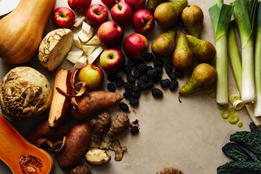 Flat lay arrangement of fresh autumn produce including butternut squash, red apples, green pears, leeks, sweet potatoes, celeriac, and blackberries on a neutral surface.