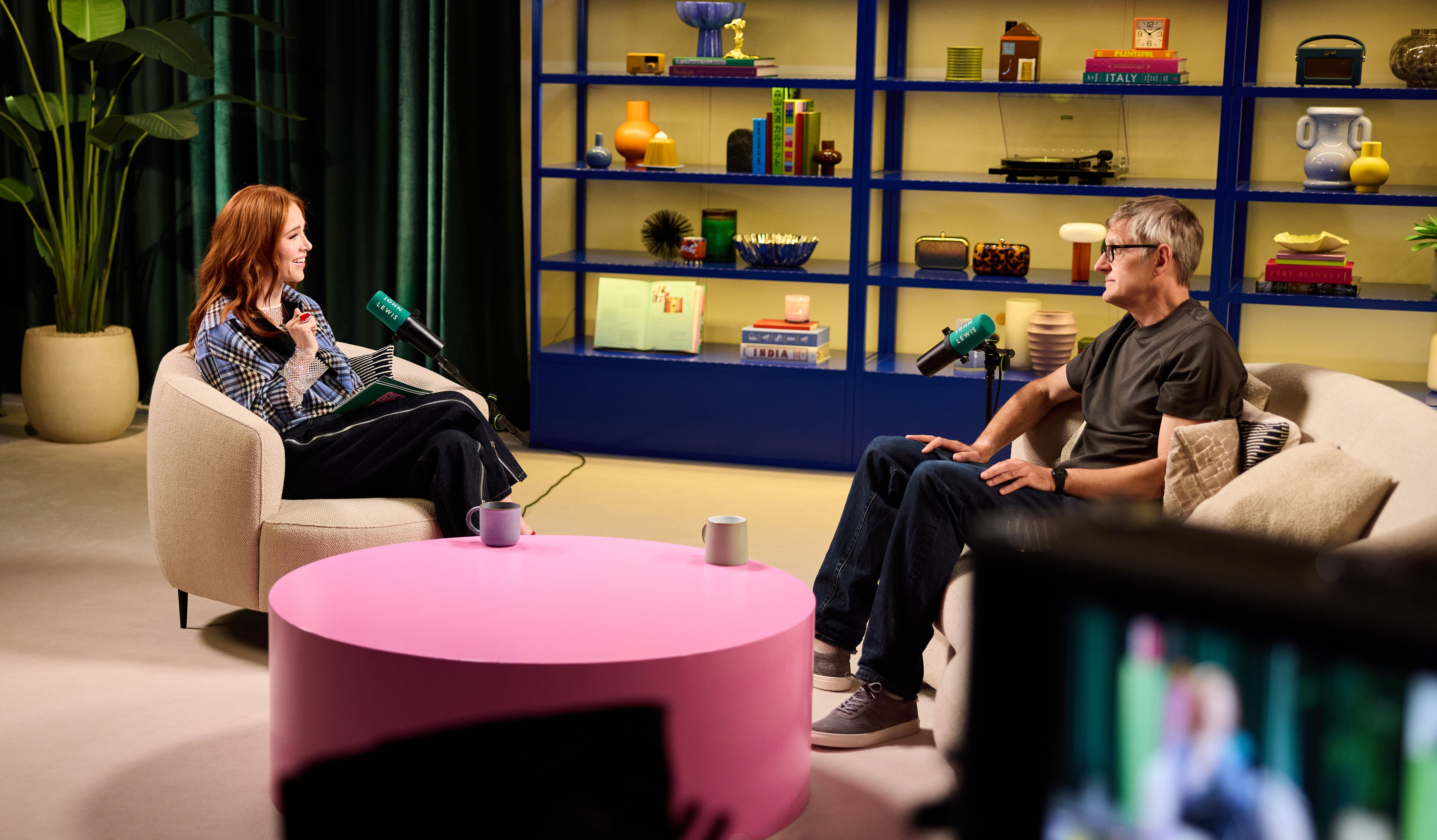 Two people sitting in a modern studio on beige and green chairs. A woman with red hair in a plaid blazer gestures with her left hand. A man with a short haircut in a black t-shirt has his hands in his lap. Large blue shelving unit holds decorative items.