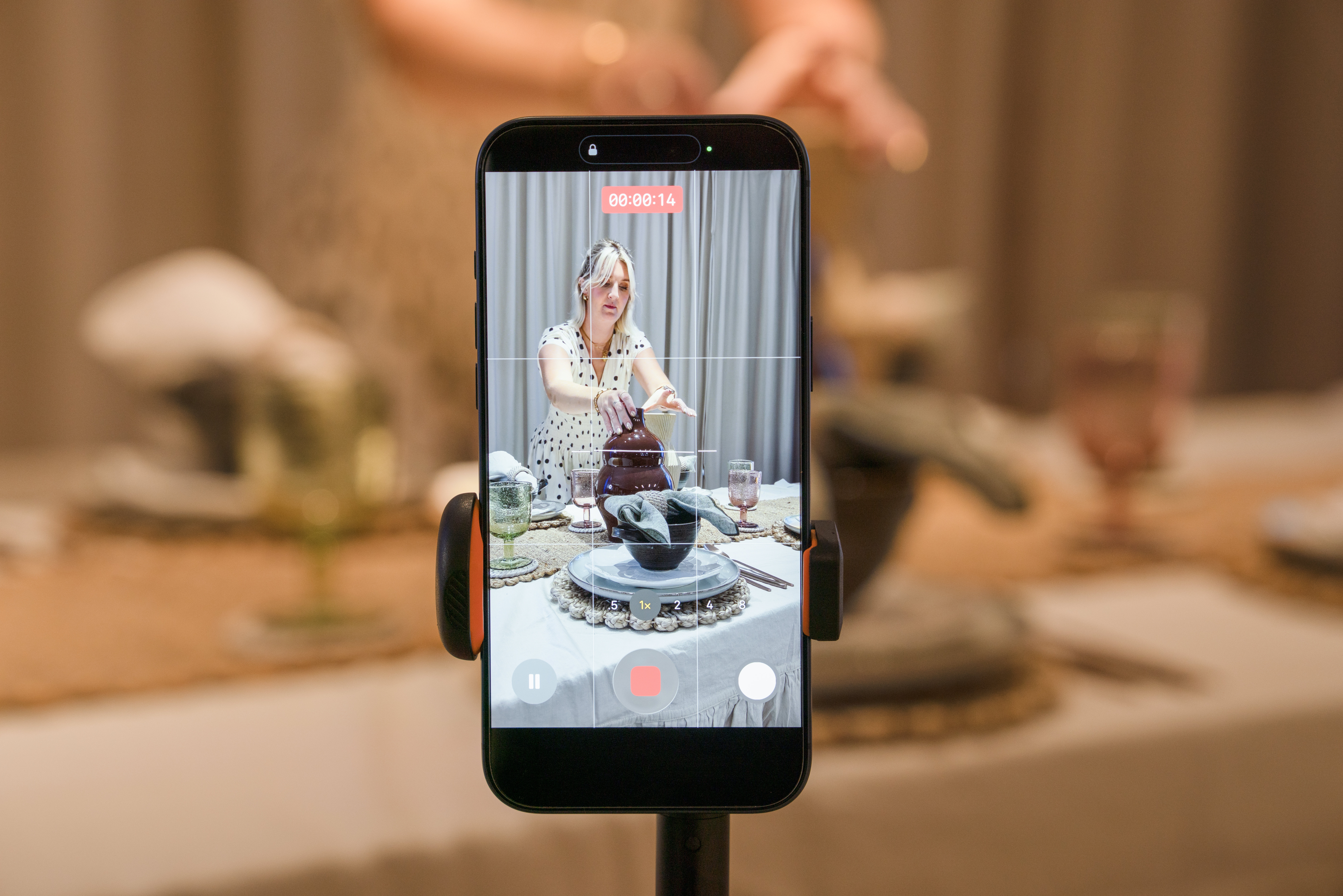 A smartphone mounted on a tripod records a video of a person setting a dining table with patterned plates, glassware, and decorative ceramics.