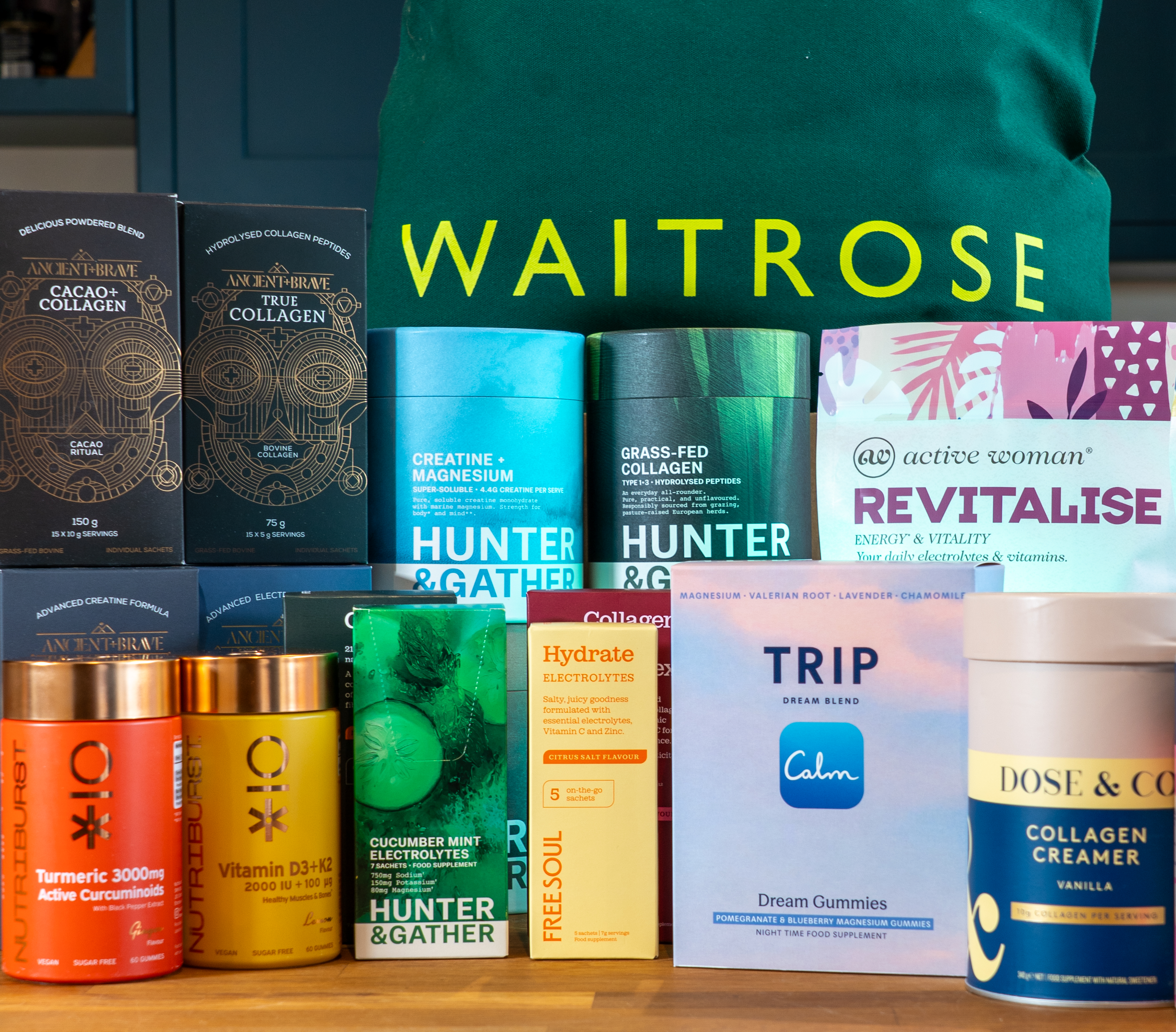 An assortment of health supplements, collagen powders, and electrolyte products displayed in front of a green Waitrose bag. Featured brands include Ancient + Brave, Hunter & Gather, Active Woman, Nutriburst, Free Soul, TRIP, and Dose & Co.