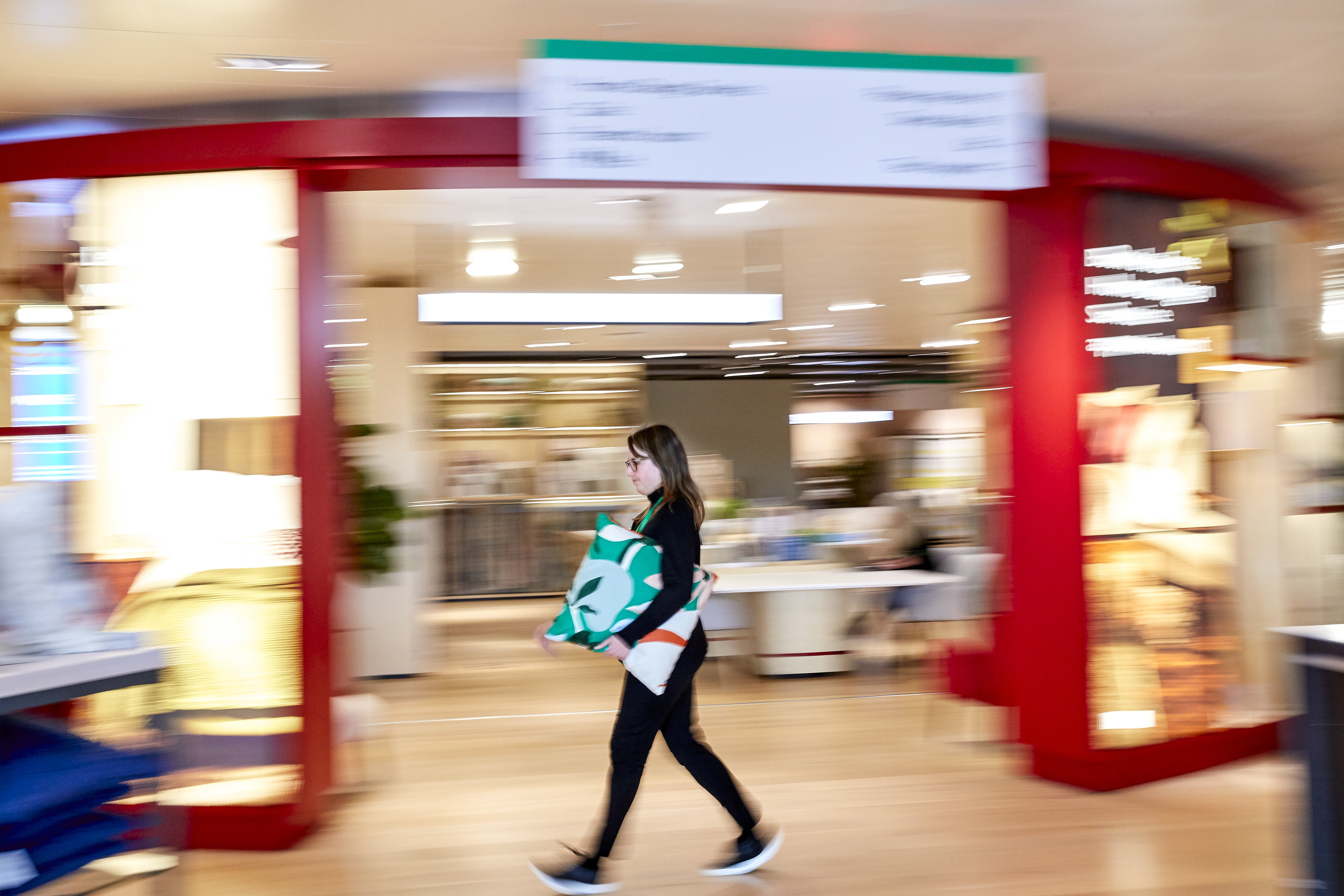 A person wearing glasses and black clothing walking through a retail store while carrying a patterned green, orange, and white cushion.