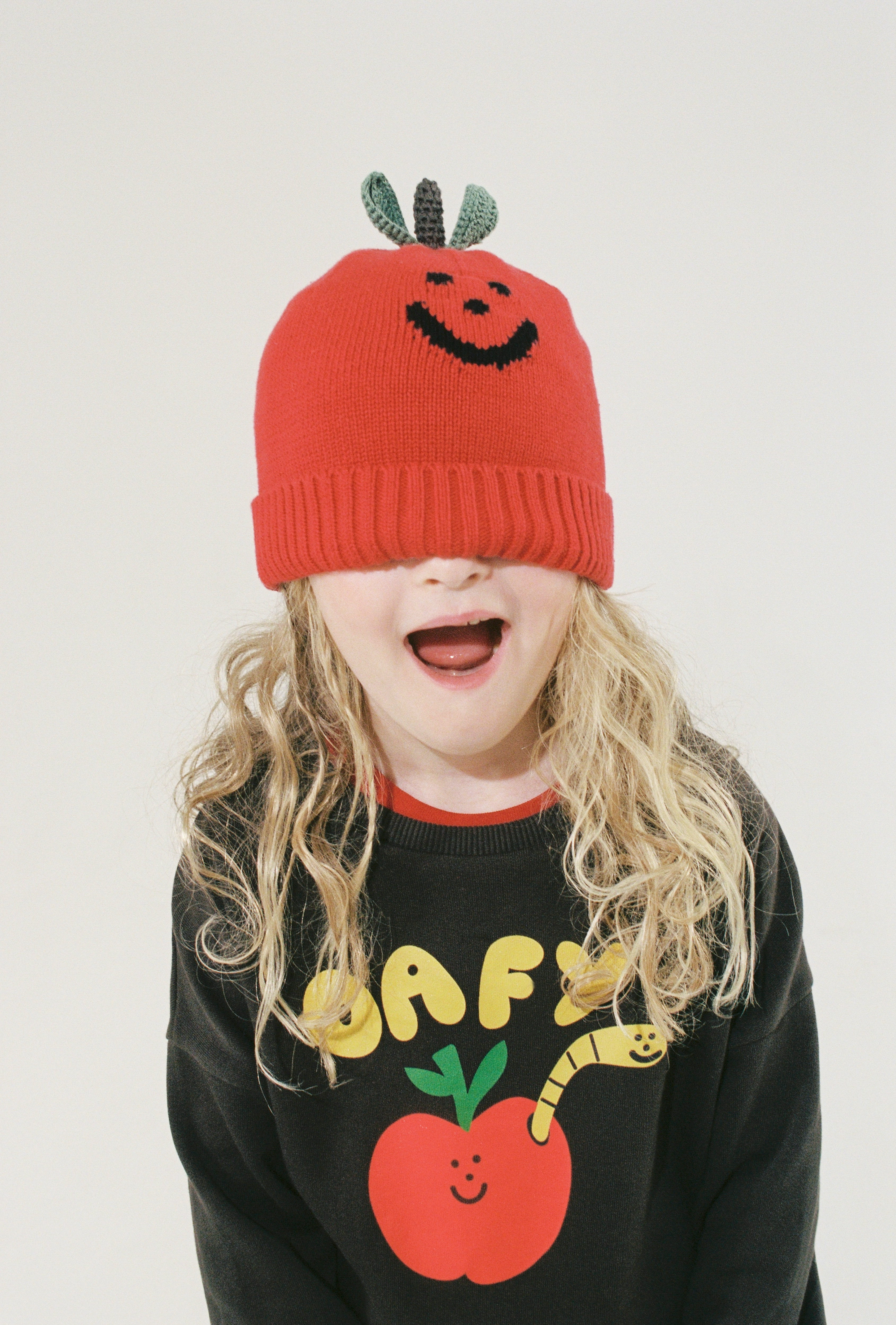 A child smiling wide with a red knitted apple beanie pulled down over their eyes, wearing a black sweatshirt with yellow text reading 