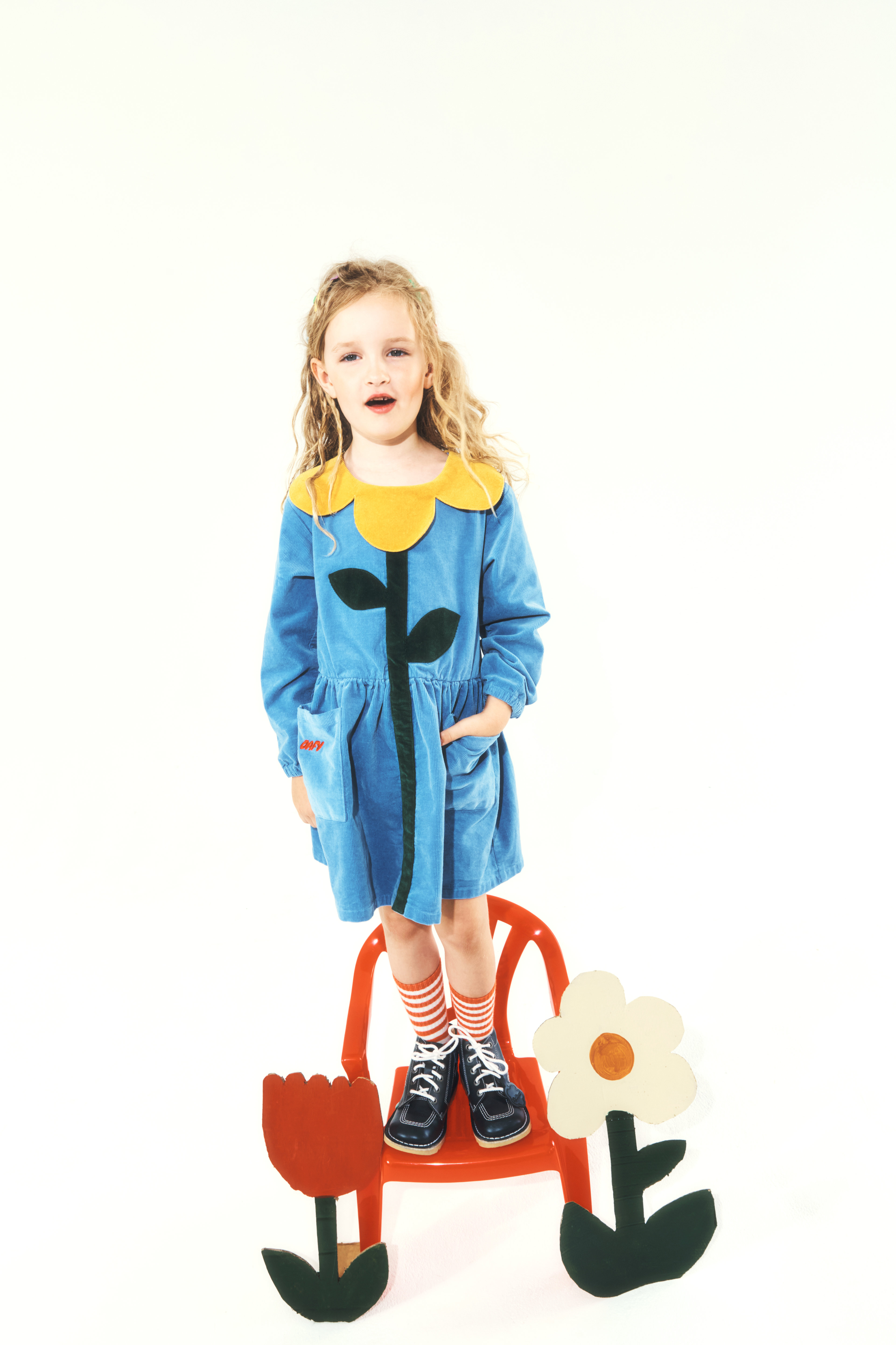 A child standing on a small red chair wearing a blue corduroy dress with a yellow flower petal collar and flower stem design, paired with striped socks and dark shoes.