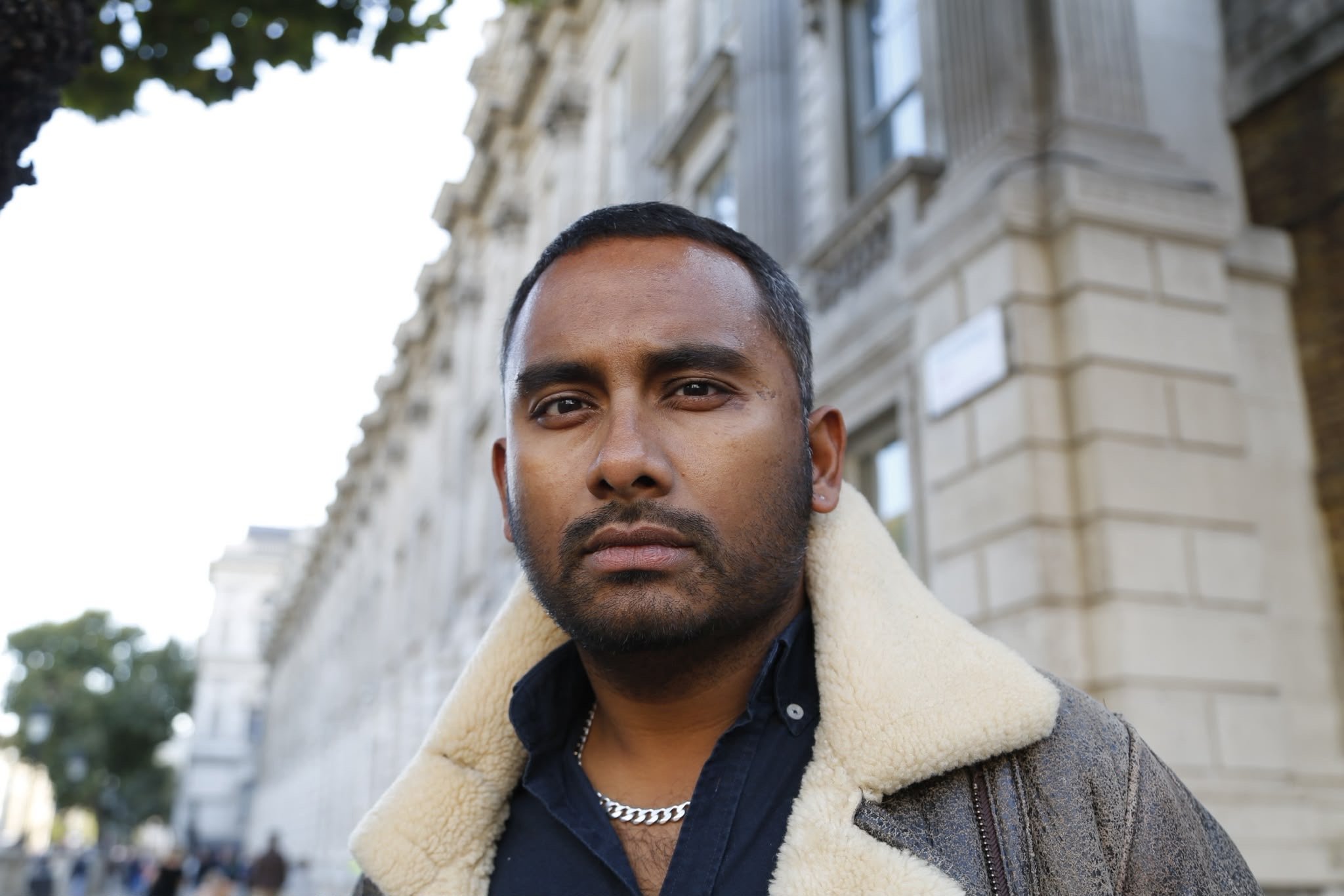 Close-up portrait of Amol Rajan wearing a shearling-lined jacket and silver chain, standing outdoors in front of a stone building.