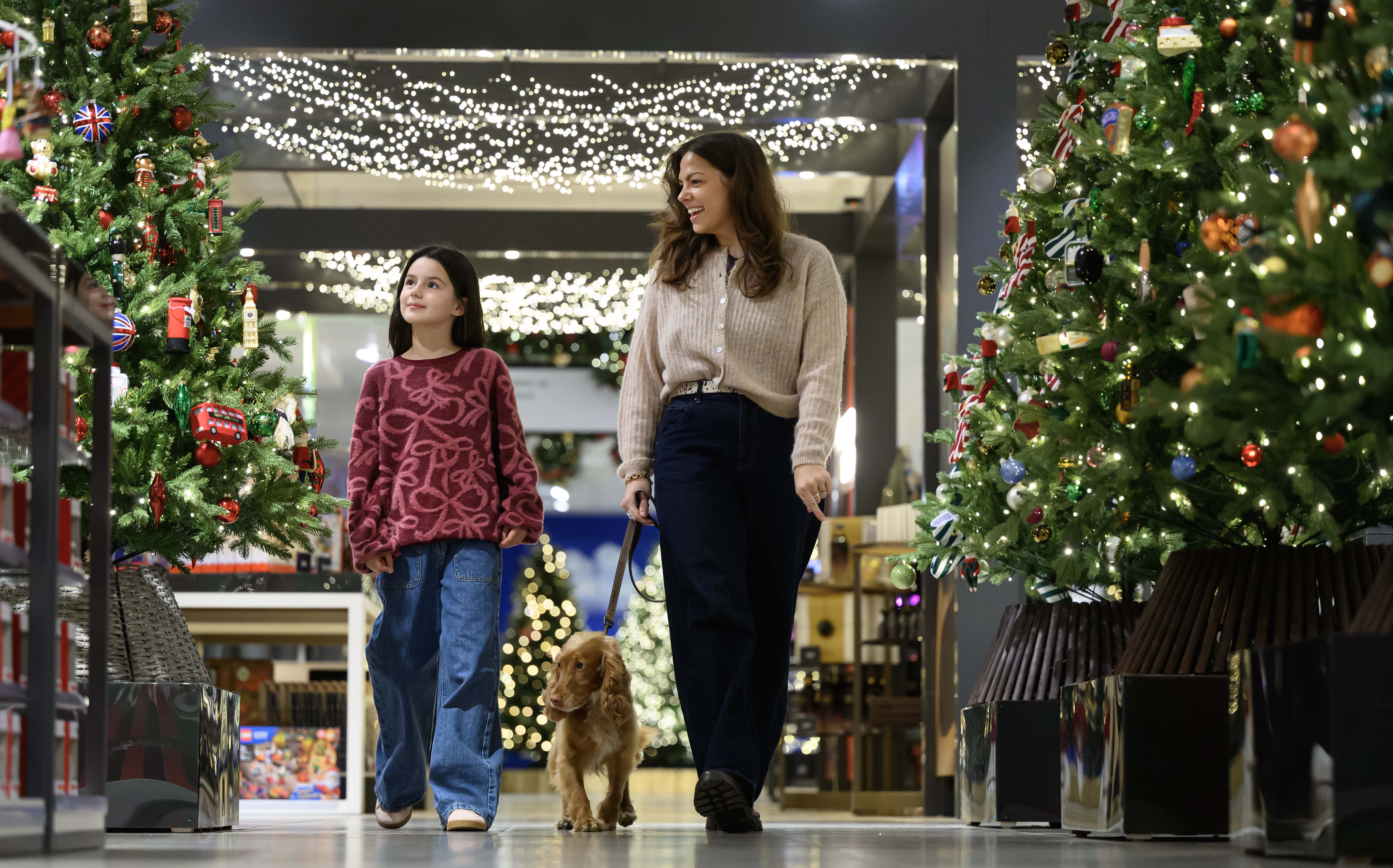 An adult and a child walking a dog on a lead through a decorated Christmas shop flanked by lit Christmas trees and overhead fairy lights.
